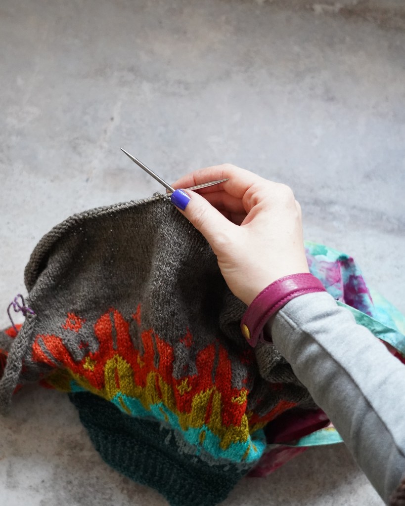 Knitting a sweater, colourful sweater in the frame also featuring a hand with blue nail polish, a grey sleeve and magenta leather bracelet.